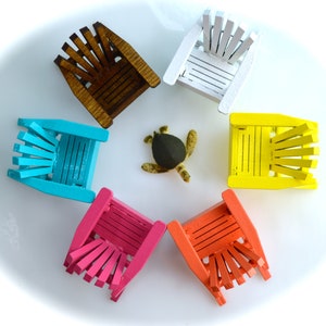 May include: A collection of miniature Adirondack chairs in a circular arrangement, each painted a different color: brown, white, yellow, orange, pink, and turquoise. A small turtle figurine is in the center. The chairs are made of wood.