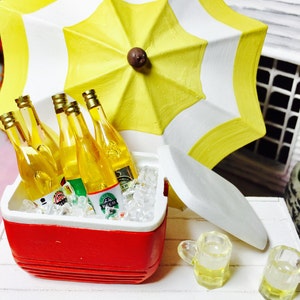 May include: Miniature scene featuring a red and white cooler filled with ice and beer bottles. A yellow and white striped umbrella provides shade. Two small beer mugs sit nearby, suggesting a summer picnic or outdoor gathering.