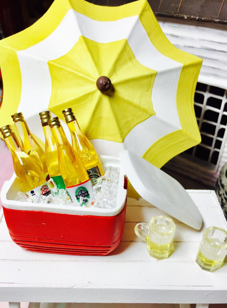 May include: Miniature scene featuring a red and white cooler filled with ice and beer bottles. A yellow and white striped umbrella provides shade. Two small beer mugs sit nearby, suggesting a summer picnic or outdoor gathering.