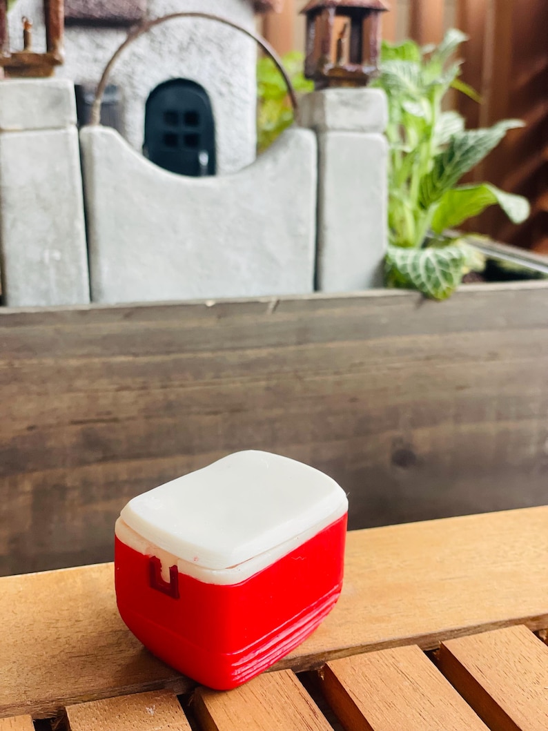 May include: A miniature red and white cooler with a white lid. The small, rectangular cooler has a red body and a white top. The cooler is sitting on a wooden surface.