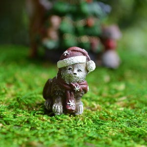 May include: A miniature figurine of a cat wearing a Christmas hat and scarf. The cat is brown with white accents and is sitting on a bed of green moss. The hat and scarf are burgundy with white snowflake details.