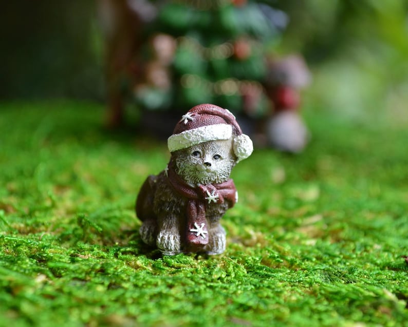 May include: A miniature figurine of a cat wearing a Christmas hat and scarf. The cat is brown with white accents and is sitting on a bed of green moss. The hat and scarf are burgundy with white snowflake details.