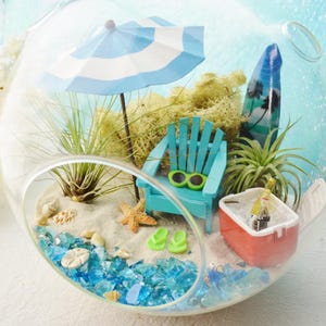 May include: A miniature beach scene inside a clear glass terrarium. It features a blue and white striped umbrella, a turquoise Adirondack chair, a surfboard, and a small cooler. The base is sand with blue glass, shells, and a starfish.