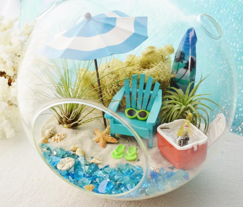 May include: A miniature beach scene inside a clear glass terrarium. It features a blue and white striped umbrella, a turquoise Adirondack chair, a surfboard, and a small cooler. The base is sand with blue glass, shells, and a starfish.