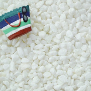 May include: A miniature beach bag figurine with a red handle and sunglasses rests on a bed of white pebbles. The bag features blue, green, and white stripes with a red base, evoking a summer theme.