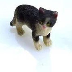 May include: A small, detailed figurine of a cat with black and white fur, standing on a white surface. The cat has pink accents on its ears and cheeks, with green eyes. The figurine is approximately 1 inch in length.