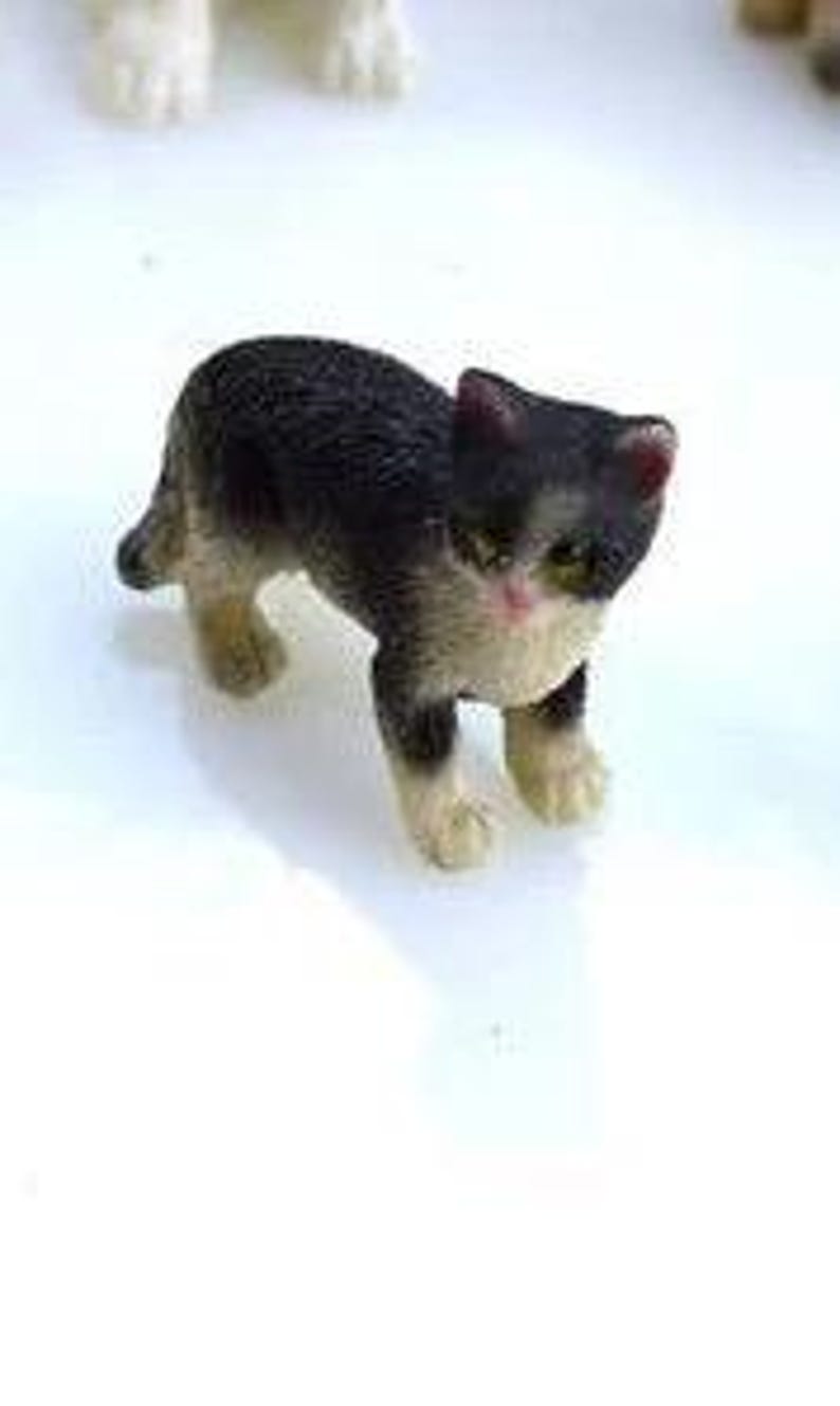 May include: A small, detailed figurine of a cat with black and white fur, standing on a white surface. The cat has pink accents on its ears and cheeks, with green eyes. The figurine is approximately 1 inch in length.
