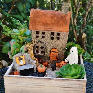 May include: A miniature Halloween-themed fairy garden scene. It features a small house with a brown roof, a wooden bench with a pillow, pumpkins, a black cat, and a white ghost figure. The scene is set in a wooden planter box with greenery.