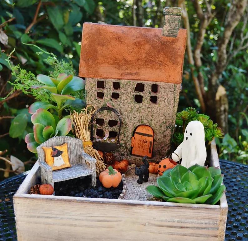 May include: A miniature Halloween-themed fairy garden scene. It features a small house with a brown roof, a wooden bench with a pillow, pumpkins, a black cat, and a white ghost figure. The scene is set in a wooden planter box with greenery.