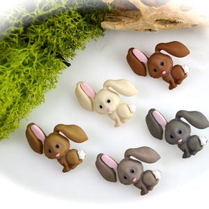 May include: A collection of small, decorative bunny figurines in various shades of brown, gray, and cream. Each bunny has long ears with pink inner ears and a fluffy white tail. The bunnies are arranged on a white surface with green moss.