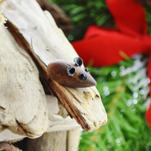May include: A wooden mouse figurine with black ears and eyes rests on a piece of driftwood. The mouse is brown with a pointed nose and a long tail. The background includes a red bow and green foliage.