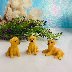 May include: Three small, golden-colored puppy figurines in various poses. One sits upright, another lies down, and the third sits with its head turned. The background includes lavender sprigs and a blurred, light blue backdrop.