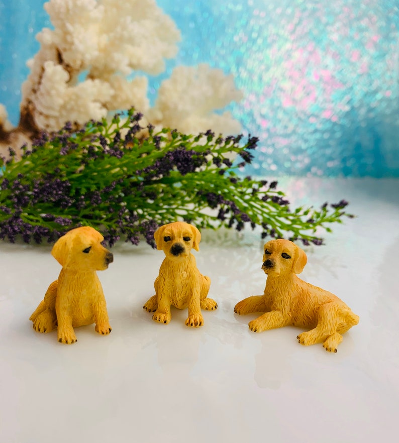 May include: Three small, golden-colored puppy figurines in various poses. One sits upright, another lies down, and the third sits with its head turned. The background includes lavender sprigs and a blurred, light blue backdrop.