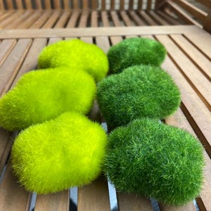 May include: Six artificial moss balls in varying shades of green and chartreuse are arranged on a wooden slatted surface. The balls have a soft, textured appearance, suggesting a natural aesthetic. The arrangement creates a visually appealing display.