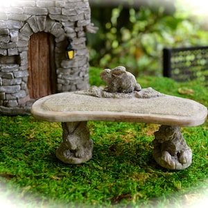 May include: A miniature stone bench with a rabbit figurine on top. The bench is light gray and has two rabbit-shaped legs. The bench is set on a bed of green moss, with a stone cottage in the background.