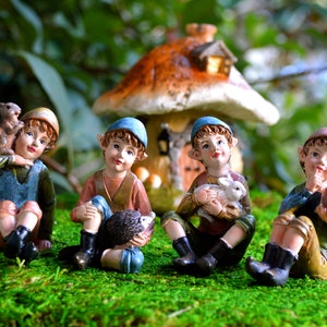 May include: Four miniature fairy figurines sitting on green moss, each holding a small animal. One holds a squirrel, another a hedgehog, a third a lamb, and the last a bird. A mushroom-shaped house is in the background.
