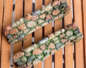 May include: Two rectangular miniature wall sections featuring a textured stone pattern with varied shades of gray, tan, and reddish-brown rocks. The gaps between the stones are filled with green moss-like detailing and thin green wire supports.