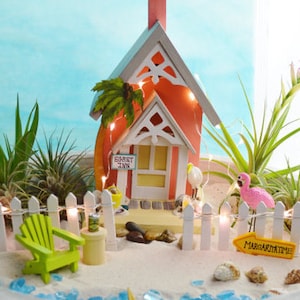 May include: A miniature beach scene with a red and white cottage, a white picket fence, and a green Adirondack chair. The cottage has a sign that reads "Egret Inn." A pink flamingo and a sign that says "Margarita Time" are also present.