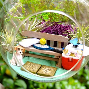 May include: A miniature terrarium scene inside a glass globe. It features a small dog figurine, a wooden bench with a tennis racket and ball, a red cooler, and decorative plants. The scene is set on a bed of light blue sand.