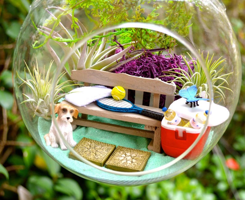 May include: A miniature terrarium scene inside a glass globe. It features a small dog figurine, a wooden bench with a tennis racket and ball, a red cooler, and decorative plants. The scene is set on a bed of light blue sand.