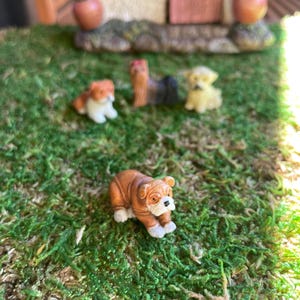 May include: Miniature dog figurines in various breeds, including a bulldog, are displayed on a bed of green moss. The bulldog is brown and white. Other dogs are tan, black, and yellow. The scene suggests a miniature garden or diorama.