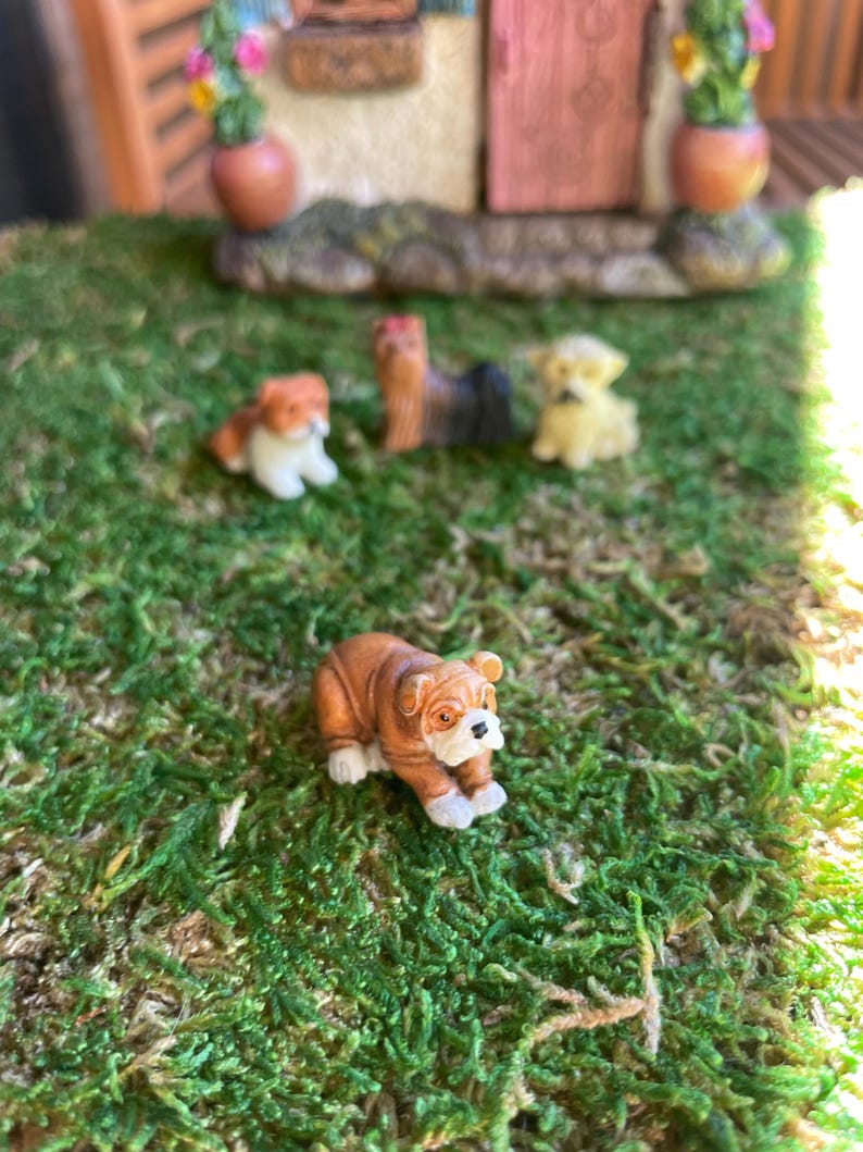 May include: Miniature dog figurines in various breeds, including a bulldog, are displayed on a bed of green moss. The bulldog is brown and white. Other dogs are tan, black, and yellow. The scene suggests a miniature garden or diorama.