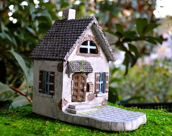 May include: A miniature fairy house with a gray tiled roof, white stucco walls, and a brown wooden door. The house has blue shutters, a small mailbox, and a stone patio. It sits on a bed of green moss.
