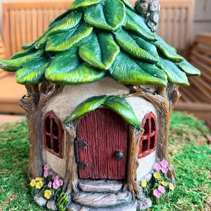 May include: A whimsical fairy house with a leaf-covered roof and a red door. The house has two arched windows with red frames and is surrounded by green moss and small flowers. The chimney is a small, textured brown structure.
