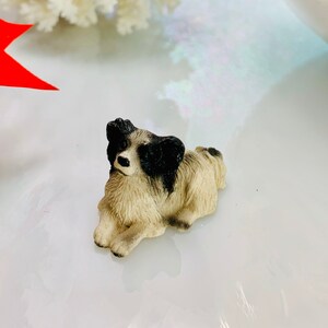 May include: A small, detailed figurine of a dog in a resting position. The dog has a cream-colored body with black markings on its head and back. The figurine is set against a bright, reflective surface.