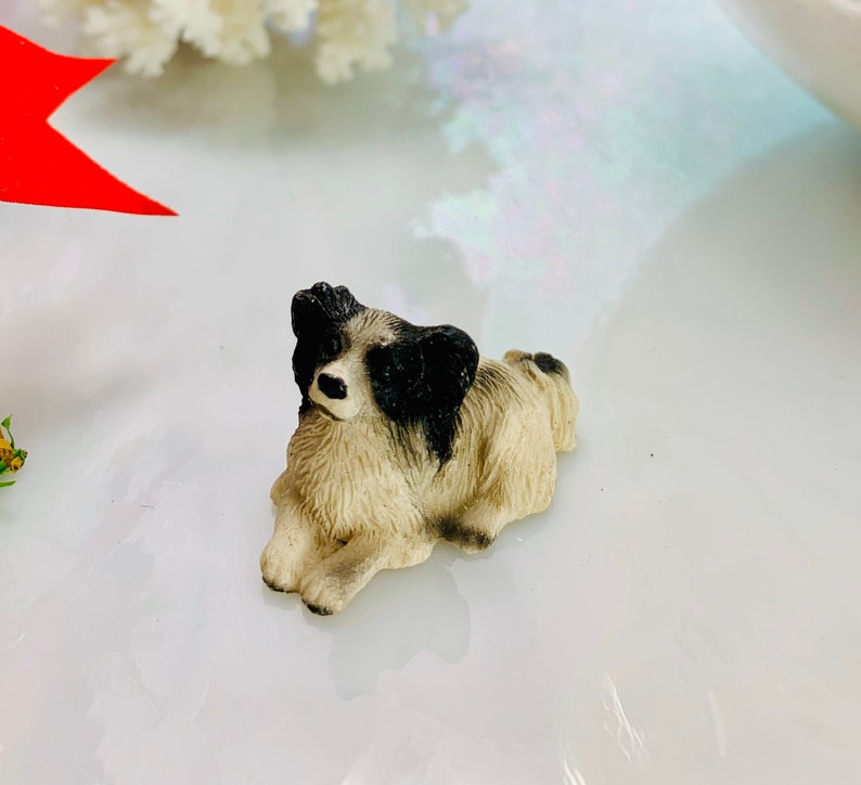 May include: A small, detailed figurine of a dog in a resting position. The dog has a cream-colored body with black markings on its head and back. The figurine is set against a bright, reflective surface.