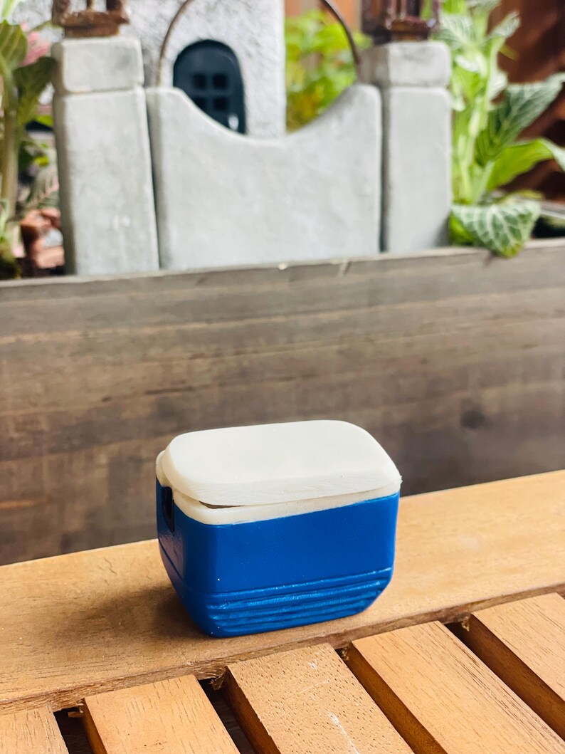 May include: A miniature blue and white cooler with a rectangular shape. The cooler has a white lid and a blue body with horizontal ridges. It is sitting on a wooden surface.