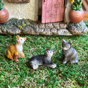 May include: Three miniature cat figurines in shades of orange, black, and gray sit on a bed of green moss. The cats are positioned in front of a miniature house with a pink door and flower pots.