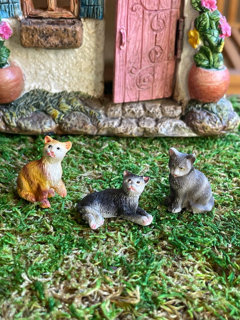 May include: Three miniature cat figurines in shades of orange, black, and gray sit on a bed of green moss. The cats are positioned in front of a miniature house with a pink door and flower pots.