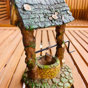 May include: A miniature wishing well figurine with a green-tiled roof, brown supports, and a stone-like base. The well features a small bucket and a decorative handle. The color palette includes shades of green, brown, and yellow.
