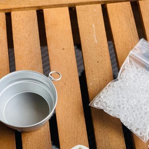 May include: A small, silver-colored metal bucket with a handle and a clear plastic bag filled with small, clear rubber bands. The items are on a wooden surface with a slatted design.