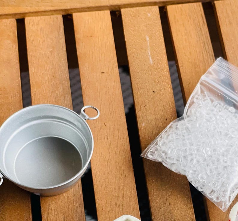 May include: A small, silver-colored metal bucket with a handle and a clear plastic bag filled with small, clear rubber bands. The items are on a wooden surface with a slatted design.