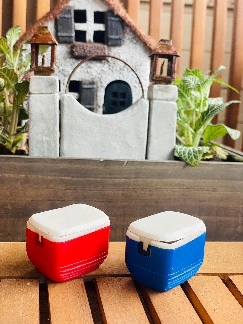 May include: Two miniature coolers, one red and one blue, each with a white lid. The coolers are rectangular with rounded edges and a handle on the side. They are set on a wooden surface, with a miniature house in the background.