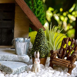 May include: A miniature garden scene featuring a rustic wooden bench, a small metal bucket, and a decorative tree. A small, white and gray bunny figurine sits on white pebbles. The scene is set against a blurred green background.