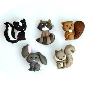 May include: A collection of five woodland animal figurines: a skunk, raccoon, beaver, bunny, and squirrel. The skunk is black and white, the raccoon has a striped tail, the beaver has a large tail, the bunny has pink ears, and the squirrel has a bushy tail.