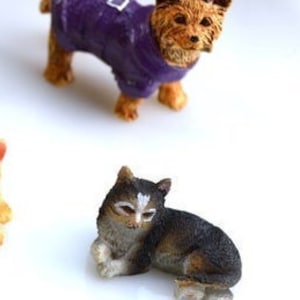 May include: Miniature figurines of a dog and a cat. The dog is brown and tan, wearing a purple coat with a silver buckle. The cat is black, brown, and white, in a resting pose.