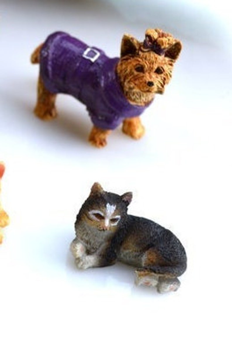 May include: Miniature figurines of a dog and a cat. The dog is brown and tan, wearing a purple coat with a silver buckle. The cat is black, brown, and white, in a resting pose.
