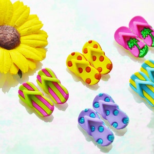 May include: A collection of colorful miniature flip-flops in various designs, including pink with green palm trees, yellow with red polka dots, and blue with yellow stripes. A large yellow sunflower is also present.