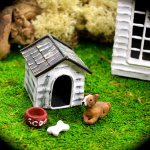 May include: Miniature dog house with a brown roof, white walls, and a black arched doorway. A small brown and white dog figurine, a red bowl, and a white bone are nearby. The scene is set on a bed of green moss.