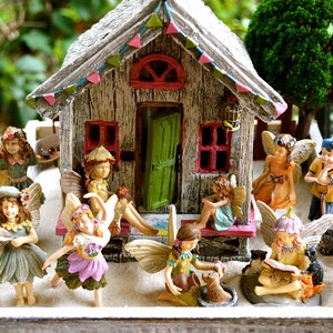 May include: A miniature fairy garden scene featuring a rustic wooden house with a green door and red-framed windows. Several fairy figurines with detailed wings and colorful outfits are arranged around the house, creating a whimsical display.