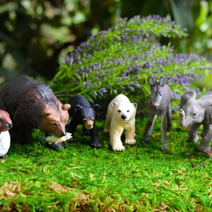 May include: A collection of miniature animal figurines, including a brown bear, black bear cub, polar bear, donkey, elephant, and fox, arranged on a bed of green moss. A brown rabbit is also present. The background features purple flowers and greenery.