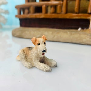 May include: A small, detailed figurine of a dog, lying down with its front paws extended. The dog has tan and white fur, with black eyes and a black nose. The figurine is set against a blurred background of a miniature building.