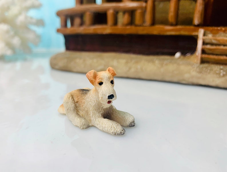May include: A small, detailed figurine of a dog, lying down with its front paws extended. The dog has tan and white fur, with black eyes and a black nose. The figurine is set against a blurred background of a miniature building.