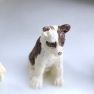 May include: A small, handcrafted dog figurine. The dog is white with brown patches on its head and back. It is sitting upright with its paws visible. The figurine has a fluffy texture and a detailed, realistic appearance.