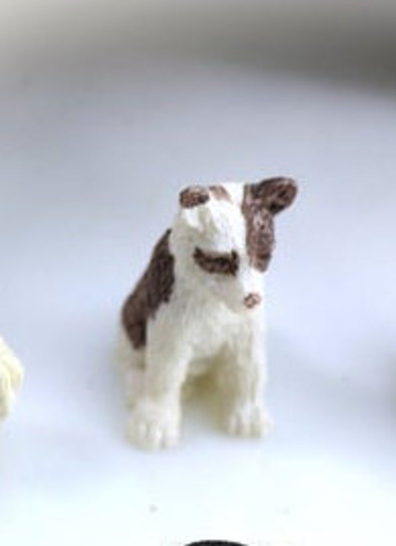 May include: A small, handcrafted dog figurine. The dog is white with brown patches on its head and back. It is sitting upright with its paws visible. The figurine has a fluffy texture and a detailed, realistic appearance.