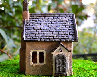 May include: A miniature brown house with a gray shingled roof, a chimney, and a small front door. The house has a window and is set on a bed of green moss, creating a whimsical fairy garden scene.